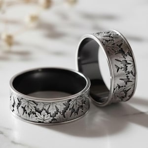 Puede incluir: Dos anillos plateados con interior negro. Los anillos presentan un detallado patrón de hojas alrededor de la banda. Se exhiben sobre una superficie de mármol blanco, con un fondo suave.