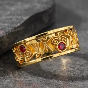 May include: A gold-colored ring with a decorative design featuring cut-out shapes and two red gemstones. The ring has an intricate pattern with a polished finish, set against a gray stone surface.