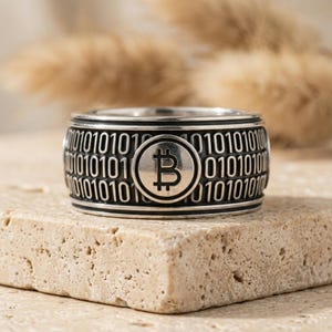 Puede incluir: Un anillo de banda ancha de plata con un patrón de código binario de ceros y unos grabado alrededor de la circunferencia, con un prominente símbolo circular de Bitcoin centrado en la parte frontal de la pieza de joyería metálica.