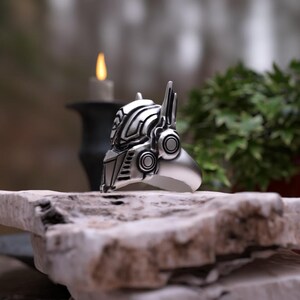 May include: A silver ring shaped like a robot's helmet, with detailed mechanical features. The ring is displayed on a rough, light-colored stone surface, with a blurred background including greenery and a candle.