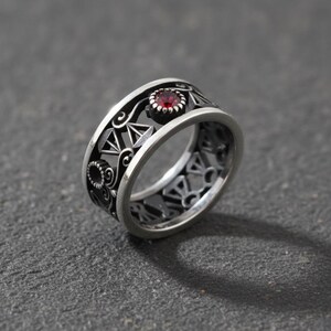 May include: A silver ring with an openwork design. The ring features a central red gemstone and intricate geometric and floral patterns. The ring is set against a dark gray background.