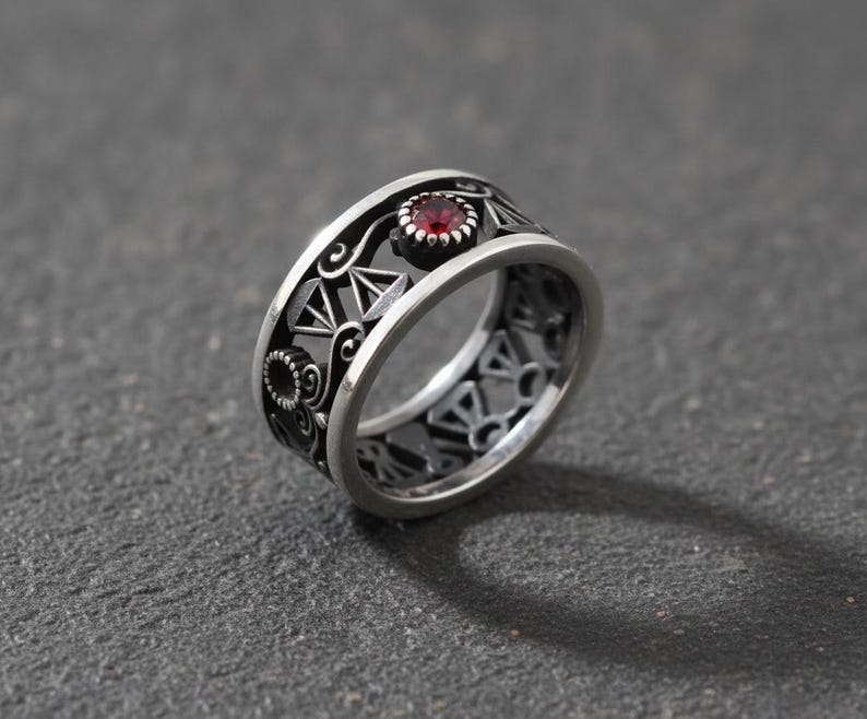 May include: A silver ring with an openwork design. The ring features a central red gemstone and intricate geometric and floral patterns. The ring is set against a dark gray background.