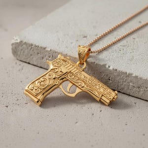 Puede incluir: Collar con colgante dorado con un diseño detallado de pistola. El colgante tiene intrincados patrones grabados y está suspendido de una delicada cadena. La joya se exhibe sobre una superficie gris claro.