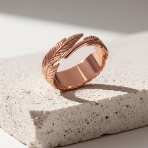 May include: Rose gold feather ring. The ring has a detailed feather design on the top, with the feather extending around the band. It is displayed on a textured, light-coloured surface. The ring's warm colour contrasts with the neutral background.