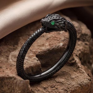 Puede incluir: Una pulsera de metal negro con forma de serpiente enroscada y un patrón de escamas texturizado a lo largo de la banda. La cabeza de la serpiente presenta una pequeña piedra preciosa redonda de color verde vibrante como ojo.