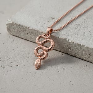 May include: Rose gold snake pendant necklace. The pendant features a detailed, coiled snake design with a textured surface. The necklace chain is also rose gold and is a delicate, linked style. The jewellery is displayed on a light grey, textured surface.
