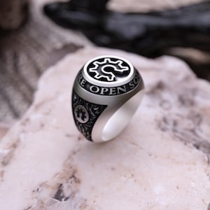 以下が含まれることがあります： 黒い歯車模様と「RE OPEN SO」の文字が刻まれたシルバーリング。明るい色の石の表面に置かれています。