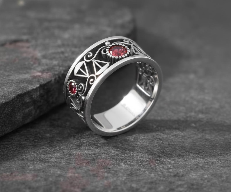 May include: A silver ring with a detailed design featuring two red gemstones. The ring has an intricate pattern with geometric shapes and swirling details. The gemstones are round and set in a bezel setting. The ring is displayed on a dark gray surface.