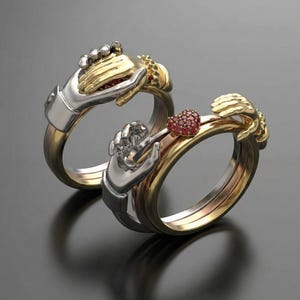Op de afbeelding: Twee in elkaar grijpende ringen met gebeeldhouwde handmotieven. De ene ring combineert zilver- en goudtinten, terwijl de andere zilveren en gouden banden heeft met een klein, rood, hartvormig element ingelegd met kleine rode steentjes.