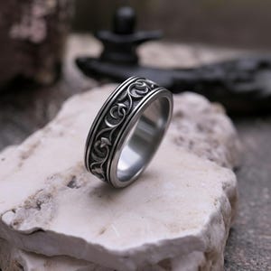 Könnte beinhalten: Ein silberfarbener Metallring mit einem durchgehenden, erhabenen Reliefmuster aus geschwungenen Ranken und Blättern auf dem äußeren Band. Die Innenseite des Rings ist glatt und poliert, während das Außendesign dunkle, vertiefte Bereiche aufweist.