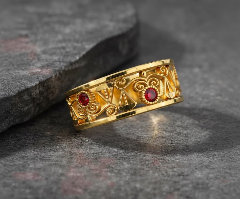 May include: A gold-colored ring with a decorative design featuring cut-out shapes and two red gemstones. The ring has an intricate pattern with a polished finish, set against a gray stone surface.