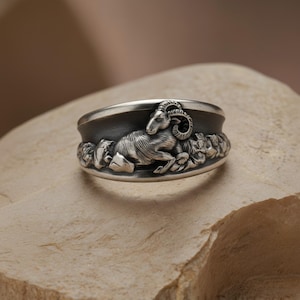 May include: A silver ring featuring a detailed relief of a ram. The ram is depicted lying down, surrounded by a textured, rocky base. The ring has a dark band and a polished silver finish, showcasing the intricate design.