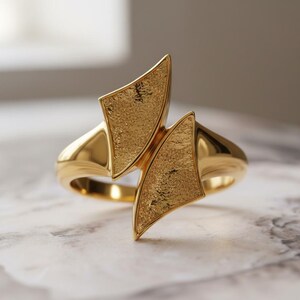Op de afbeelding: Een gouden ring met een uniek ontwerp. De ring heeft twee getextureerde, ruitvormige elementen die van elkaar af staan. De band is glad en gepolijst, wat een contrast creëert met de getextureerde details.