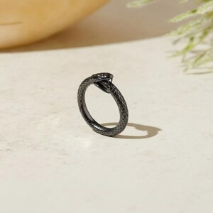 Könnte beinhalten: Ein schwarzer Ring in Form einer gewundenen Schlange mit einer strukturierten Oberfläche, die Schuppen nachahmt. Der Schlangenkopf befindet sich oben am Ringband, wobei das Maul leicht geöffnet ist, was ein durchgehendes kreisförmiges Design ergibt.