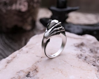 May include: A silver ring featuring a skeletal hand design holding a faceted gemstone. The ring is set on a light-colored stone surface, with a blurred background of plants and dark objects.