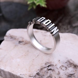 May include: A silver ring featuring the word "ODIN" in raised, block letters. The ring is displayed on a light-coloured, textured stone, with a blurred background of foliage and a dark surface.