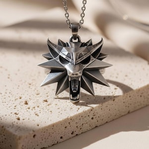 May include: Silver-toned pendant necklace featuring a stylized wolf head with sharp teeth and pointed ears. The pendant is surrounded by a starburst design. The necklace is displayed on a textured, light-colored surface.