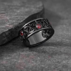 May include: A black ring with a detailed design featuring scales and red gemstones. The ring has a wide band and a polished finish, set against a textured gray surface.