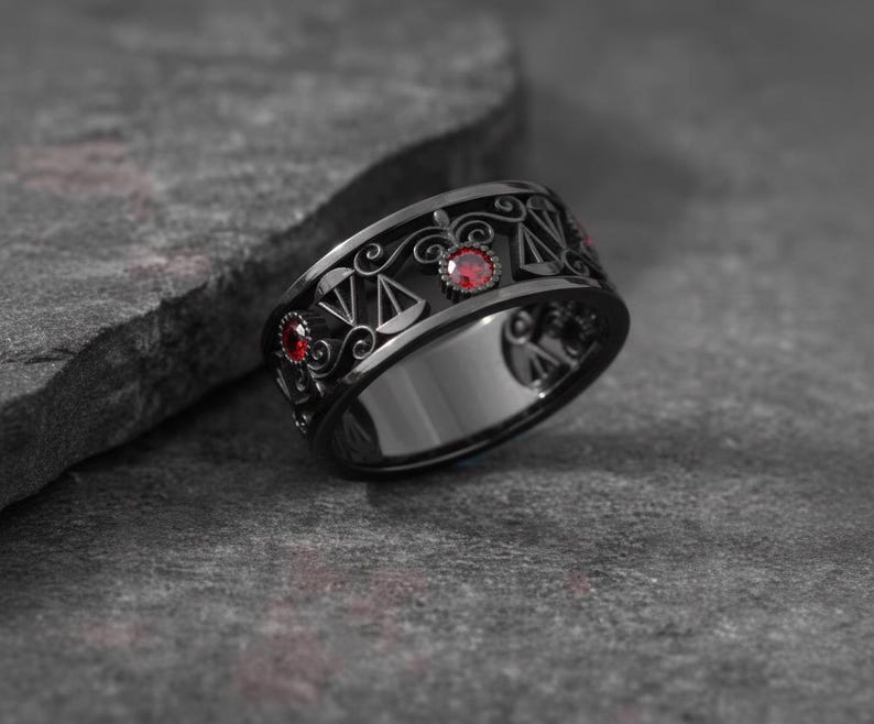 May include: A black ring with a detailed design featuring scales and red gemstones. The ring has a wide band and a polished finish, set against a textured gray surface.