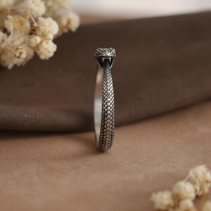 May include: A silver ring featuring a detailed snake head design at the top. The band of the ring is textured with a repeating scale pattern that wraps around the finger. The metal has an oxidized finish to highlight the intricate snake scales.