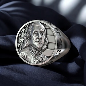 Könnte beinhalten: Ein silberner Ring mit einer runden Oberfläche, die ein Porträt von Benjamin Franklin vor dem Hintergrund der amerikanischen Flagge zeigt. Der Ring ist mit dem Wort "Freiheit" beschriftet.
