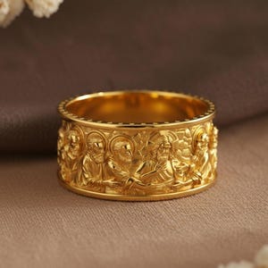 Pode incluir: Um anel dourado com entalhes em relevo detalhados de figuras. O anel tem uma faixa larga com um design contínuo ao redor da circunferência. O anel é exibido em uma superfície de tons neutros.