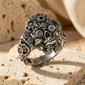 May include: A silver-toned ring featuring a detailed skull design integrated with numerous mechanical gears and cogs. The surface of the ring displays a complex arrangement of interconnected circular gear components and two curved tubes extending from the jaw.