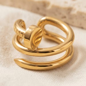 May include: A gold-toned ring shaped like a coiled nail, featuring a smooth, polished finish. The design includes a rounded nail head at the top and a tapered point at the bottom, with small, subtle indentations along the curved metal band.