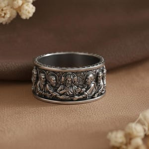 Könnte beinhalten: Ein breiter Ring aus silberfarbenem Metall mit einem detaillierten Relief des Abendmahls, das sich um das gesamte Band zieht. Das Design zeigt Figuren mit Heiligenscheinen vor einem steinartigen Hintergrund mit einer dunklen Oxidationspatina.