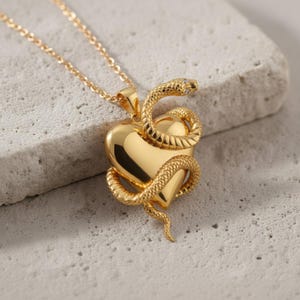 May include: A gold necklace with a heart-shaped pendant and a snake design. The snake is coiled around the heart, and the necklace has a fine chain. The pendant is highly polished, reflecting light.