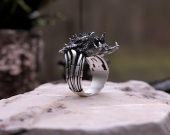 May include: A silver ring with a detailed, textured creature design. The ring features a wide band and a three-dimensional sculpture of a mythical beast. The ring is displayed on a light-coloured stone surface.