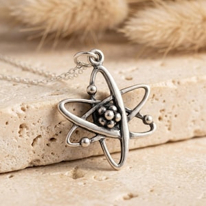 Op de afbeelding: Een zilverkleurige metalen hanger in de vorm van een atoom met een centrale groep kleine bolletjes omringd door drie elkaar kruisende elliptische banen. De hanger is bevestigd aan een fijne zilverkleurige schakelketting.