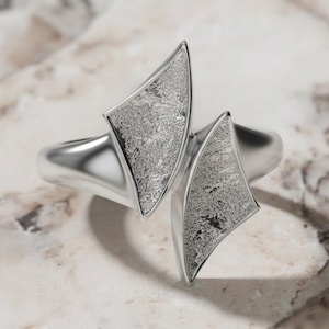 May include: A silver-toned bracelet with two asymmetrical, textured, kite-shaped accents. The bracelet has a polished, smooth band and is displayed on a light-colored, marbled surface. The accents have a detailed, speckled pattern.