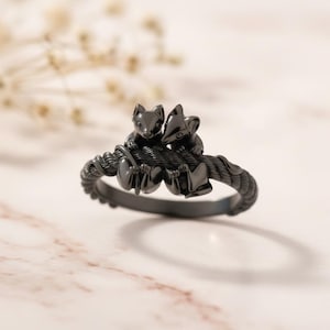 Könnte beinhalten: Ein schwarzer Ring mit zwei stilisierten Tierfiguren, die auf einem Seil sitzen. Der Ring hat eine matte Oberfläche und liegt auf einer Marmoroberfläche. Das Design des Rings ist detailliert und aufwendig, mit Fokus auf die Tierfiguren.