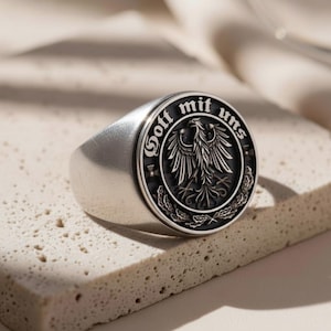 May include: A silver-toned signet ring featuring a circular face with a black recessed background. The center displays an embossed eagle emblem surrounded by the text "Gott mit uns" in a gothic font, with oak leaf detailing at the bottom.
