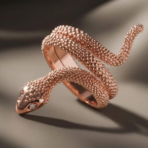 Puede incluir: Un anillo de serpiente en oro rosa con una superficie texturizada similar a escamas que se enrolla alrededor del dedo. La cabeza de la serpiente tiene dos pequeñas piedras redondas y transparentes como ojos, con un diseño en espiral.