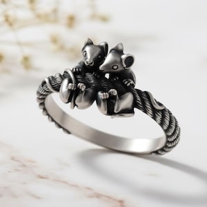 Könnte beinhalten: Ein silberner Ring mit zwei detaillierten Mäusefiguren, die auf einem seilartigen Band sitzen. Der Ring hat ein strukturiertes Design und eine polierte Oberfläche, die ein skurriles Tiermotiv zeigt.