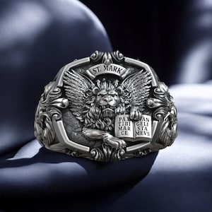 May include: A silver ring with an intricate design featuring a winged lion holding an open book with the text "Pax tibi Mar ce" and "Van geli sta mevs". The ring is inscribed with "St. Mark".