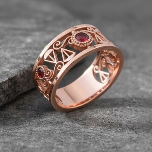 May include: Rose gold ring with intricate filigree design and two red gemstones. The ring features openwork patterns and geometric shapes, showcasing a blend of elegance and artistry. The gemstones are round and set in a bezel setting.