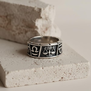 Könnte beinhalten: Ein silberfarbener Ring mit dem Sternzeichen Waage. Der Ring hat ein Banddesign mit den Waagschalen und anderen astrologischen Symbolen. Der Ring wird auf einer strukturierten, hellfarbigen Oberfläche präsentiert.