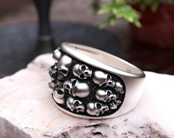 May include: A silver-toned ring featuring a wide band decorated with multiple small, raised skull motifs. The skulls are arranged in a cluster across the front surface of the band, set against a dark, recessed background to highlight the details.