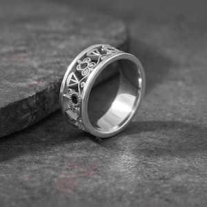 May include: A silver ring with an intricate openwork design featuring geometric shapes and floral patterns. The ring has a wide band and a polished finish, reflecting light. The ring is displayed on a dark gray surface.