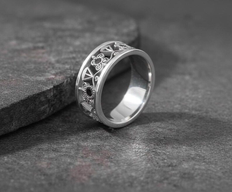 May include: A silver ring with an intricate openwork design featuring geometric shapes and floral patterns. The ring has a wide band and a polished finish, reflecting light. The ring is displayed on a dark gray surface.