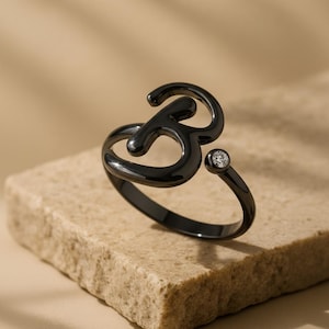 May include: A black ring shaped like the letter "B", featuring a small, clear gemstone. The ring is displayed on a light brown, textured surface. The design is modern and minimalist, suitable for everyday wear.