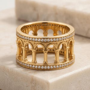 May include: A gold ring featuring an architectural design with a series of vertical columns and arches. The top and bottom edges of the band are lined with small, clear, round-cut stones that encircle the entire circumference of the ring.