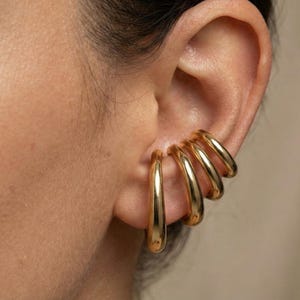 May include: Close-up of a person's ear adorned with multiple gold-colored ear cuffs. The ear cuffs are smooth, curved, and have a polished finish. The jewelry adds a touch of elegance and style.