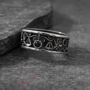 May include: A silver ring with a black interior and intricate designs. The ring features scales, circles, and other decorative elements. The ring is set against a textured gray surface.