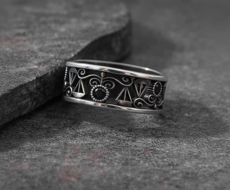 May include: A silver ring with a black interior and intricate designs. The ring features scales, circles, and other decorative elements. The ring is set against a textured gray surface.