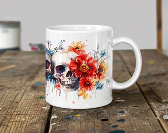 Op de afbeelding: Witte keramische mok met een aquarelontwerp van een schedel versierd met bloemen. De bloemen zijn rood, oranje, geel en blauw. De schedel is wit met zwarte ogen en een zwarte neus.