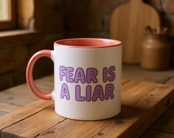 Puede incluir: Taza de cerámica blanca con interior y asa rosas. La taza presenta el texto "FEAR IS A LIAR" en letras moradas. La taza está sobre una tabla de cortar de madera.